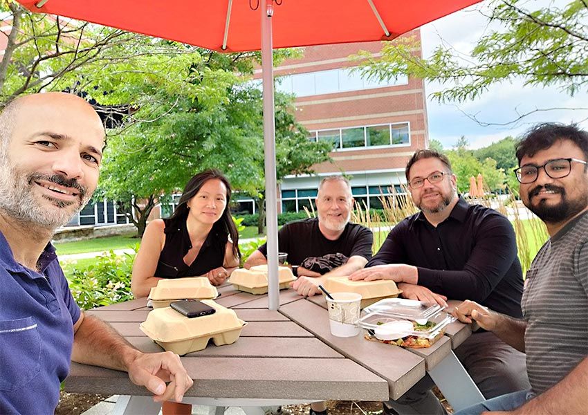 Five people seated at a picnic table with an orange umbrella and trees in the background. 