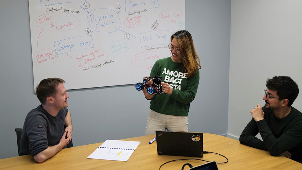 Brenda stands at a table in front of a whiteboard and holds a wheeled device. On the table are a marker, laptop, and open notebook. Two men seated at the table look at the device.