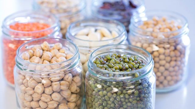 Multiple clear glass jars containing different types of dried legumes and pulses, including chickpeas, green mung beans, red lentils, and other beans, arranged in a group on a white surface.