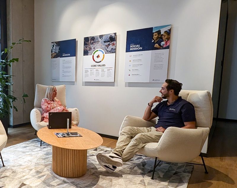A male and female are seated in front of a low oval table in an office environment and having a conversation.