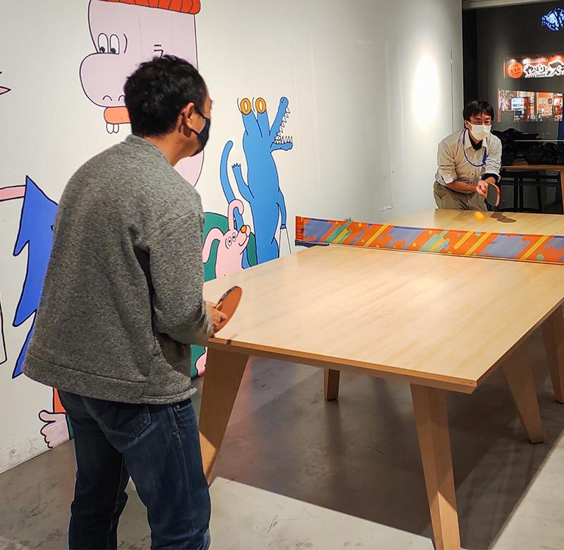 Two people wearing masks and playing table tennis in a rec room in the office.