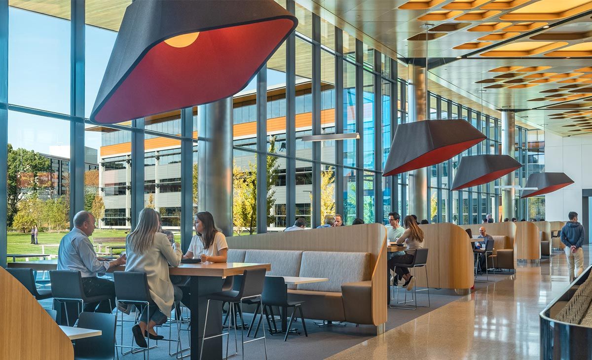 Bright, sunny cafeteria space with large windows and modern overhead lighting and design elements. Some tables are occupied by people having lunch. 