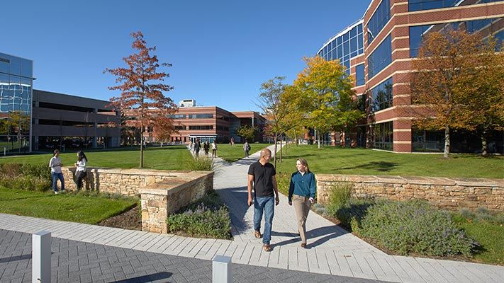 People walking on a sidewalk in a grassy quad surrounded by brick and glass buildings.