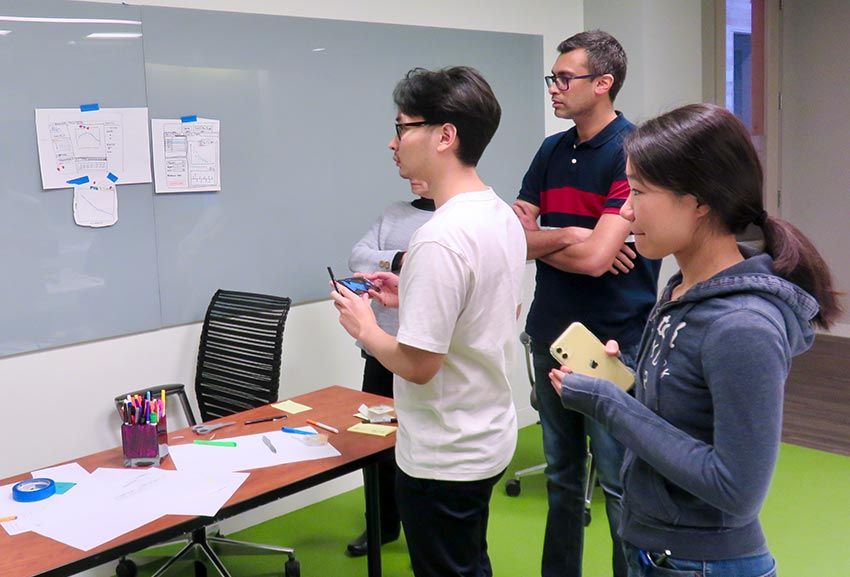 Qifang and her cross-functional product team standing in front of a white board assessing design ideas. 