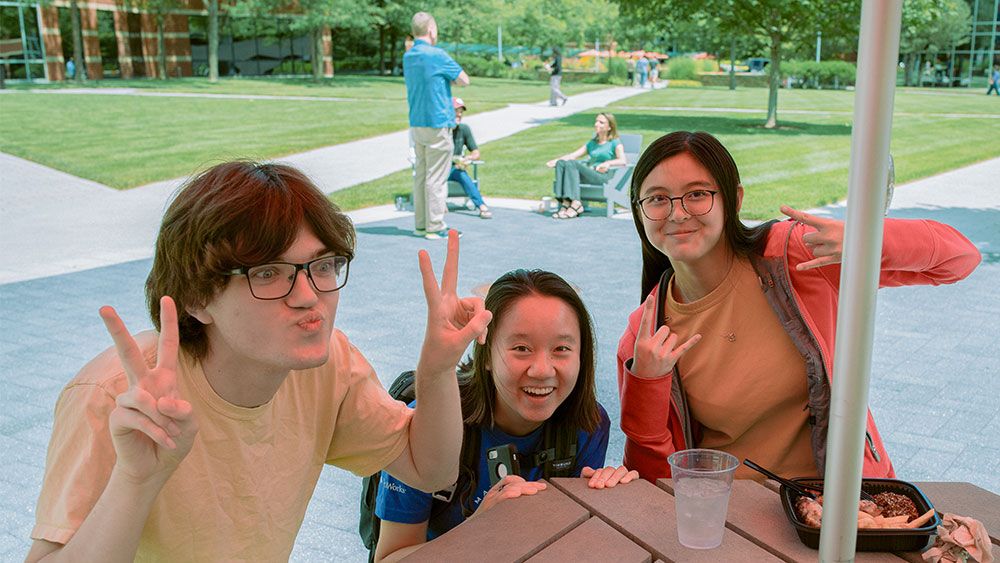 Three interns making silly faces. They are having lunch at an outdoor table with a green space and other staff enjoying a sunny day in the background.