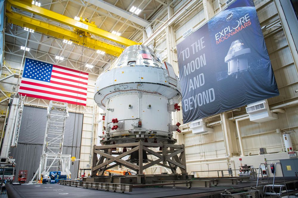 Das Orion-Raumfahrzeug in einer großen Anlage mit der Flagge der Vereinigten Staaten an einer Wand und einem Banner mit der Aufschrift „To the Moon and Beyond“ an einer anderen Wand.