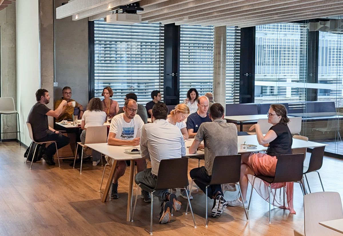 Several workers are seated at two side-by-side tables, interacting with each other, looking at material in front of them, or drinking from cups.