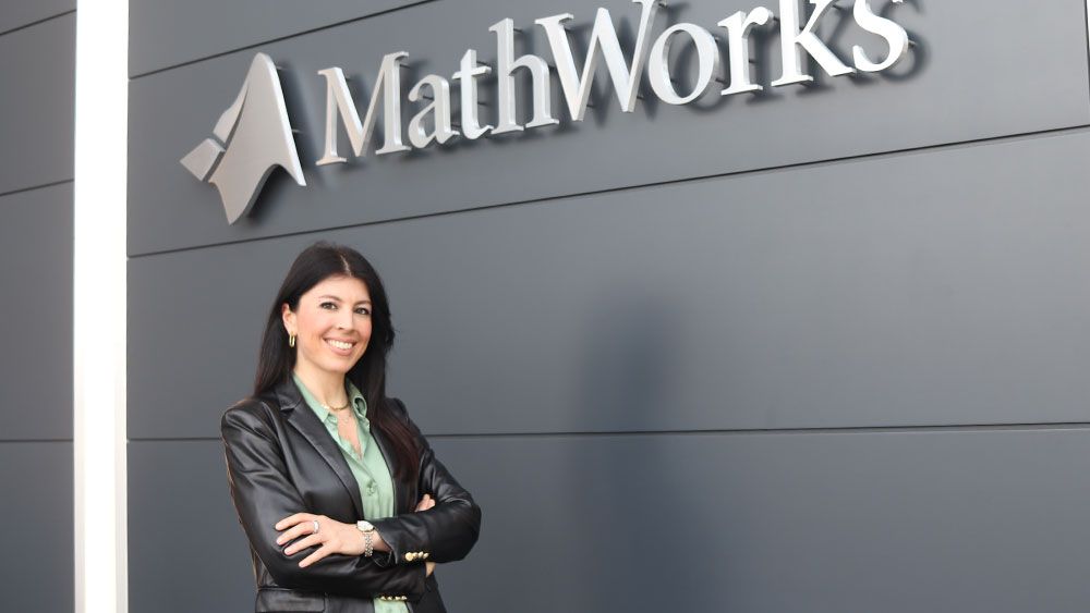 Smiling woman standing with arms crossed in front of gray interior wall with MathWorks sign.