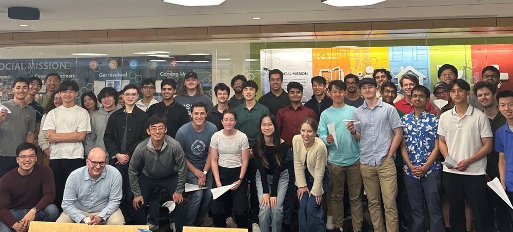 A large group of people smiling and posing together indoors in front of a colorful wall. Some hold paper airplanes.