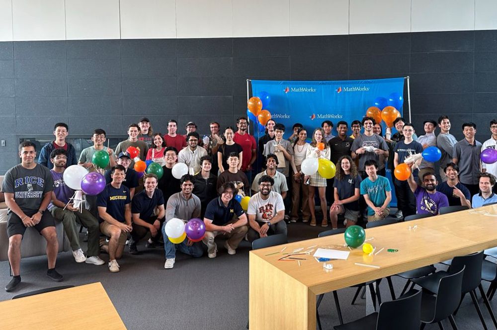 A group of people hold colorful balloons and pose in front of a MathWorks banner in a large room.
